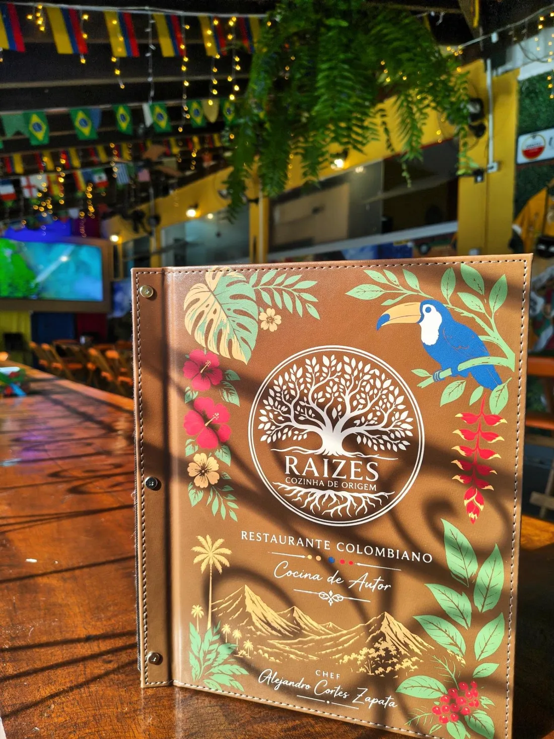Cardápio de couro da Raízes em pé sobre a mesa de madeira, com tucano e flores na capa; ao fundo, a varanda com bandeirinhas, luzinhas e samambaias